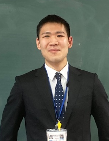 写真:黒板を背に立つスーツ姿の男性